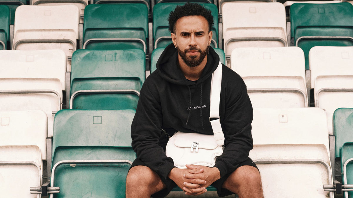 A man with a beard sits in a stadium seating area wearing a black hoodie, black shorts, and a white crossbody bag with white straps. The background consists of rows of green and white stadium seats.