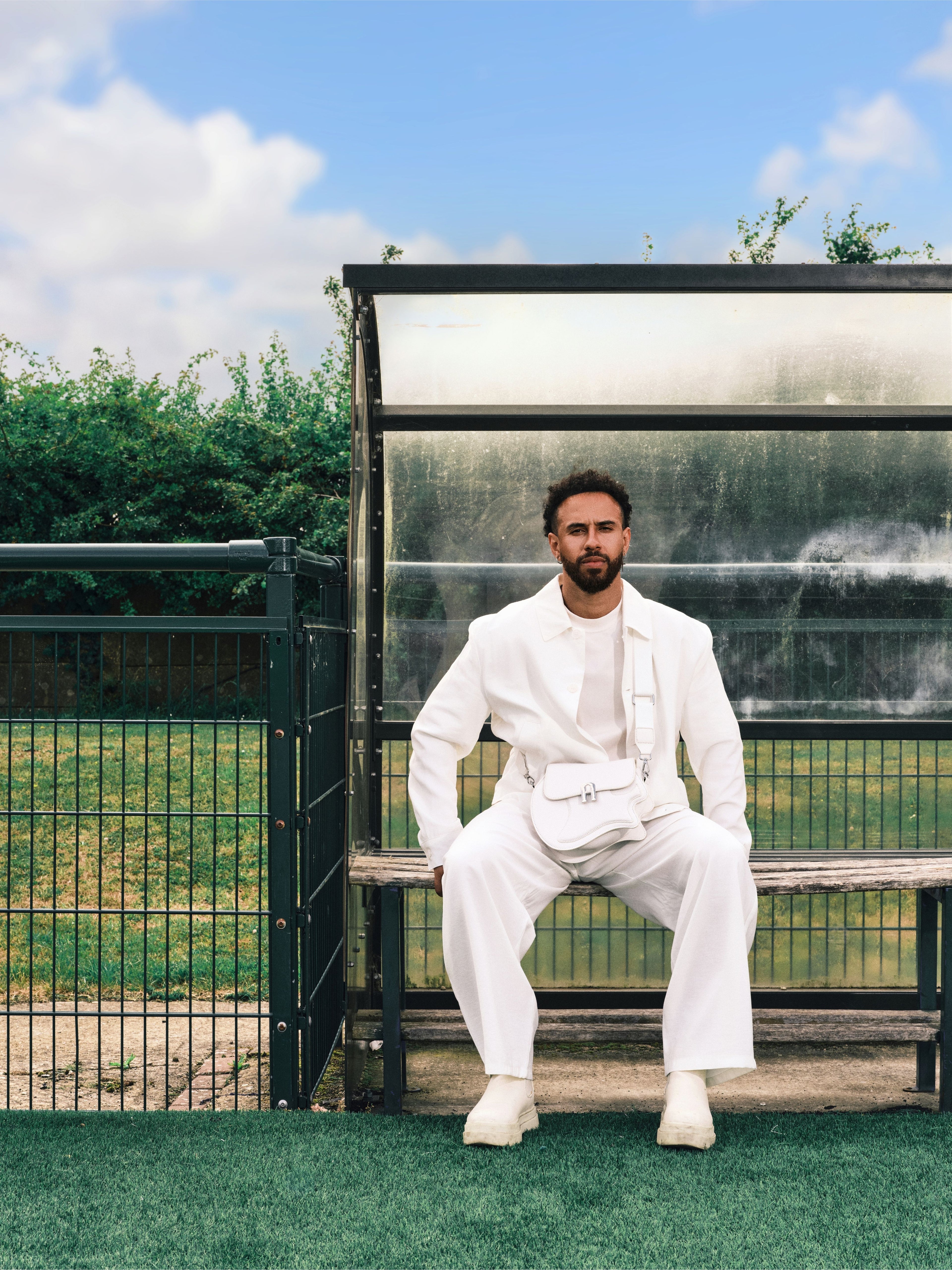 Sam Cox wearing a white three-piece suit and white sneakers sits on a bench in front of a glass shelter outdoors on a green lawn under a blue sky.
